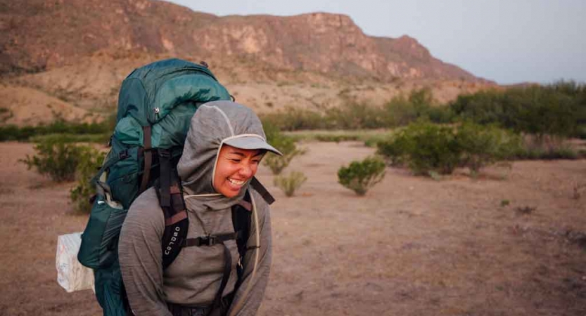 a woman wearing a backpack laughs in front of a mountainous desert landscape